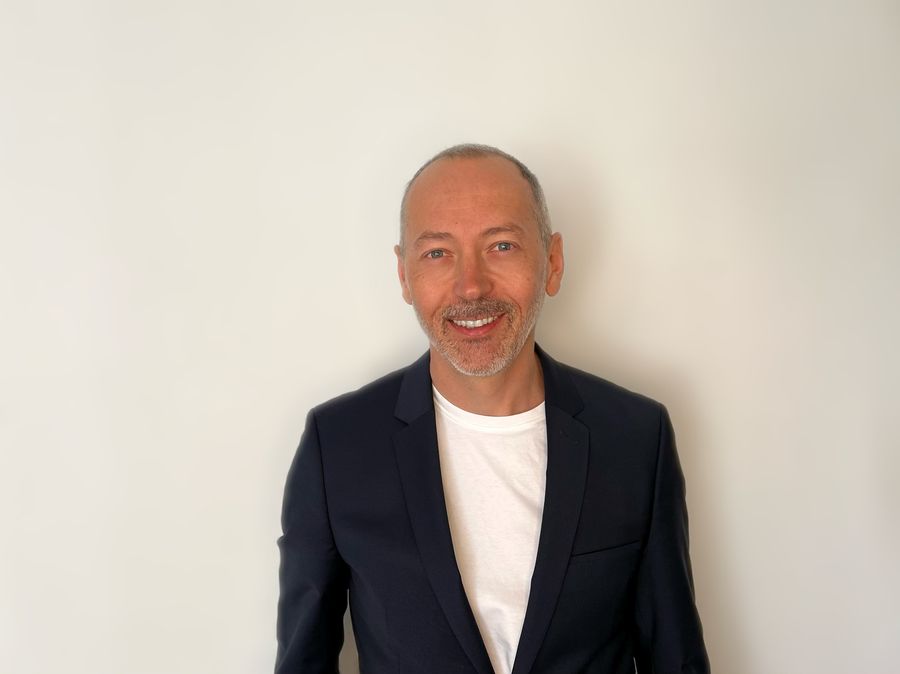 Krisztián Galánthai Fekete, smiling, in a dark blazer and white T-shirt against a light wall.